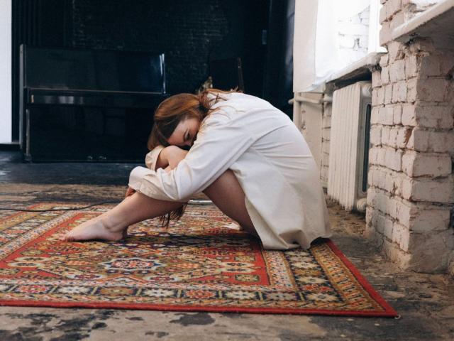 FreyaDak, with brown hair, is crouched on a patterned rug, wearing a white dress, her ass down, arms wrapped around her knees, in a dimly lit room.