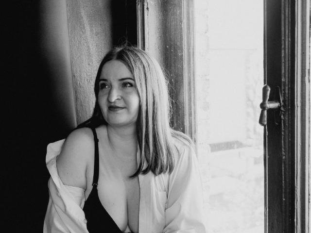 Black and white photo of FreyaDak with medium-length blonde hair, wearing a black bra and open white shirt, standing by a window, showing her tits, smiling softly.