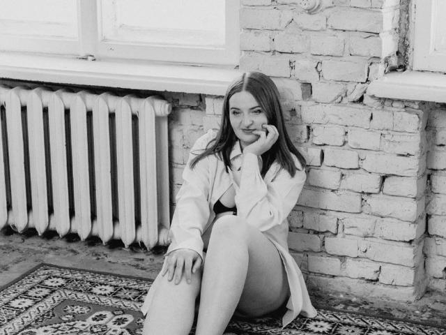 FreyaDak con i capelli lunghi, seduta contro un muro di mattoni, indossa una camicia aperta che lascia scoperte le tette e biancheria intima nera, appoggia il mento sulla mano e sorride. Foto in bianco e nero..