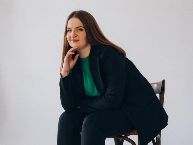 FreyaDak with long brown hair, green top, black jacket, and black pants, sitting on a wooden chair, hand near chin, slight smile, white background.