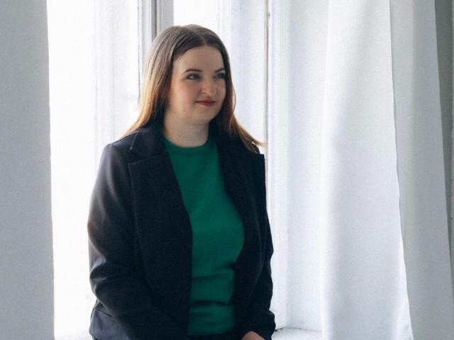 FreyaDak has brown hair, green sweater, black blazer, and red lipstick. She's standing in front of white curtains, smiling slightly. Her tits are small, and she's cute.