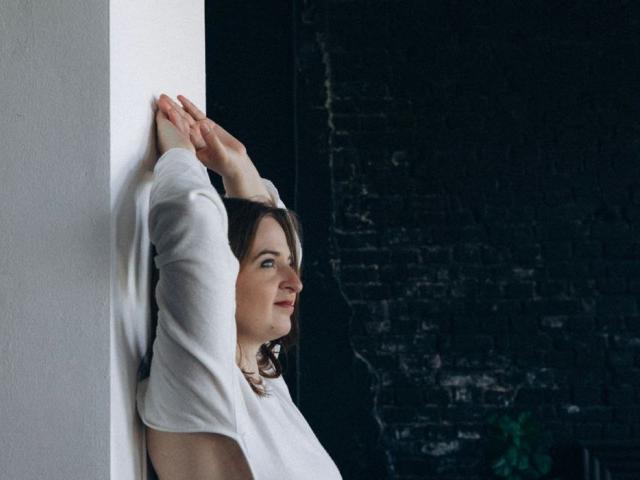 FreyaDak leans against a white wall, arms raised, wearing a white long-sleeve top, with a subtle smile, dark brick wall in the background.