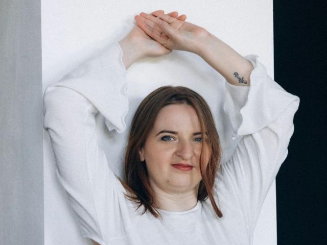 FreyaDak tiene los brazos levantados, mostrando un tatuaje en la muñeca derecha, y lleva una camisa blanca de manga larga, con cabello castaño y una ligera sonrisa..