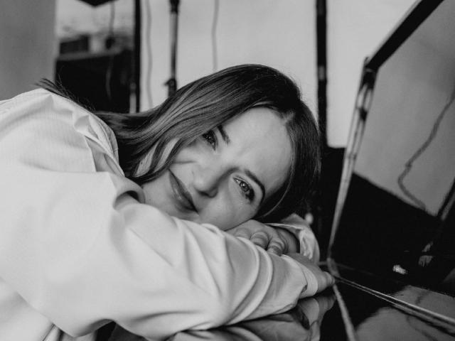 FreyaDak with her head resting on a laptop, wearing a white shirt, her eyes looking slightly upwards, with long, straight hair, in a black-and-white image.