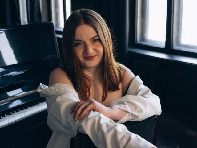 FreyaDak, a cute woman with brown hair and blue eyes, sits at a piano, wearing a white shirt off her shoulders, exposing her boobs, smiling gently at the camera.