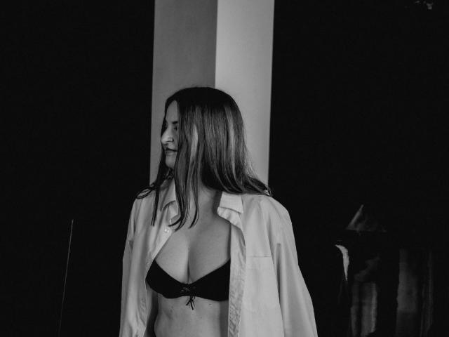 Fotografía en blanco y negro de FreyaDak con cabello largo, sonriendo, vistiendo una camisa abierta y un sujetador negro, mostrando sus pechos, de pie en un entorno oscuro y minimalista..