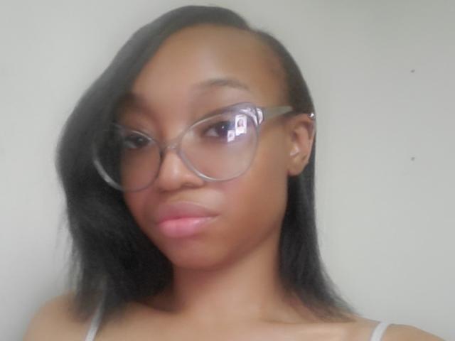 NadiaaSexy with dark skin, black shoulder-length hair, and wearing clear glasses, has a small nose and full pink lips, looking slightly to the side with a neutral expression.