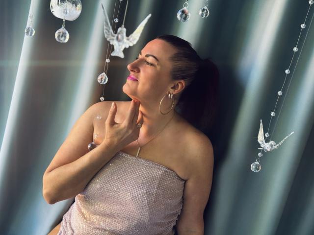 DianaMGG with pink lipstick, eyes closed, wearing a sparkly strapless dress, touching her chin, hoop earrings, brown ponytail, hanging crystal decorations, soft light, relaxed expression.