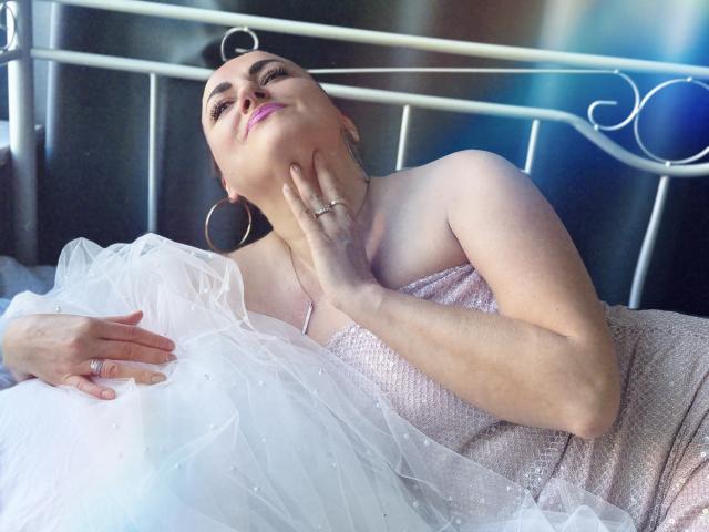 DianaMGG with pink lipstick, lying on bed, wearing a sparkly pink dress and white tulle, hand on neck, gold hoop earrings, ring on finger, smiling slightly.