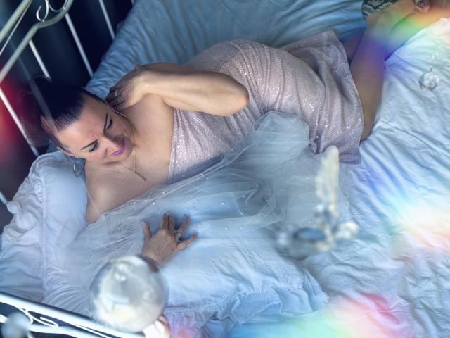 DianaMGG, com os seios quase descobertos por um vestido rosa brilhante, deitada em uma cama branca, com as mãos no peito, com uma expressão de angústia, com efeitos de luz em tons de arco-íris..