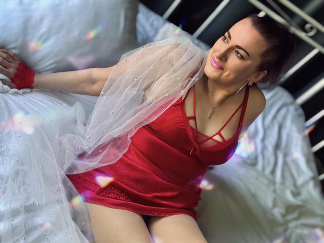 DianaMGG in a red satin dress with crisscross straps, sitting on a bed, white sheer fabric draped over her shoulder, red wristband, looking at camera with a smile.
