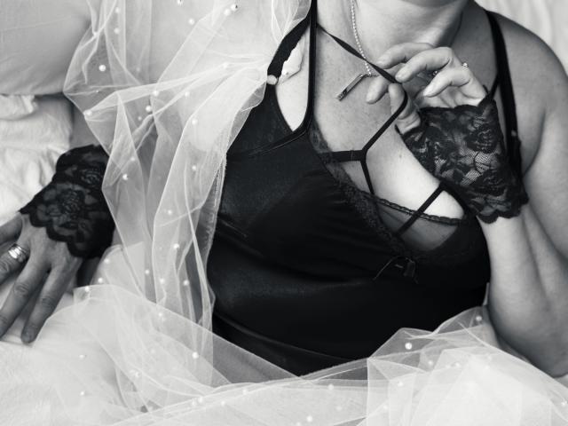 DianaMGG's tits in a black lace bra, wearing lace finger gloves and a sheer, dotted, white veil, holding glasses, with a ring on her finger. Black-and-white photo.
