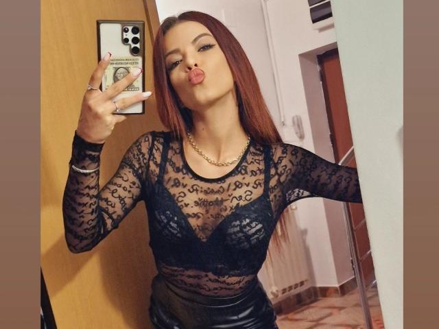 RinaQueenie with long red hair, pouting lips, wearing a black lace top, showing her tits, holding a phone, and standing in a room with a wooden door.