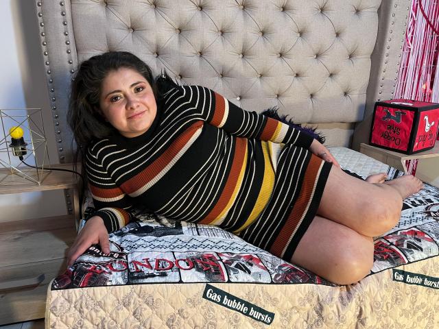 TiffanyAmazing, a plus-sized woman with dark hair, wears a striped black, orange, and yellow dress, lying on a bed with a patterned quilt, her ass and thighs visible.