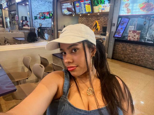 SiennaTaylor with dark hair, red lipstick, wearing a white cap, denim dress, and gold necklace, showing her tits, in a fast food restaurant, taking a selfie.