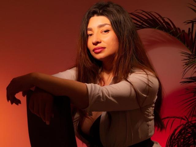Michonnek with long brown hair, pink lipstick, and a slight smile, wearing a white top, arms resting on her knees, looking confident, red and orange lighting, potted plant.