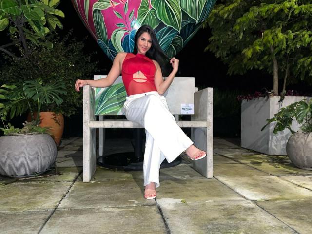 AmaiaCooper with long black hair, red cut-out top, white pants, and red sandals, sitting on a wooden bench, holding her hair, with her bare ass slightly visible.