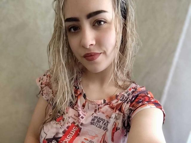 EleanorEustacia has wet blonde hair, wearing a colorful, ruffled shirt, smiling with red lipstick, showing her tits in a selfie.