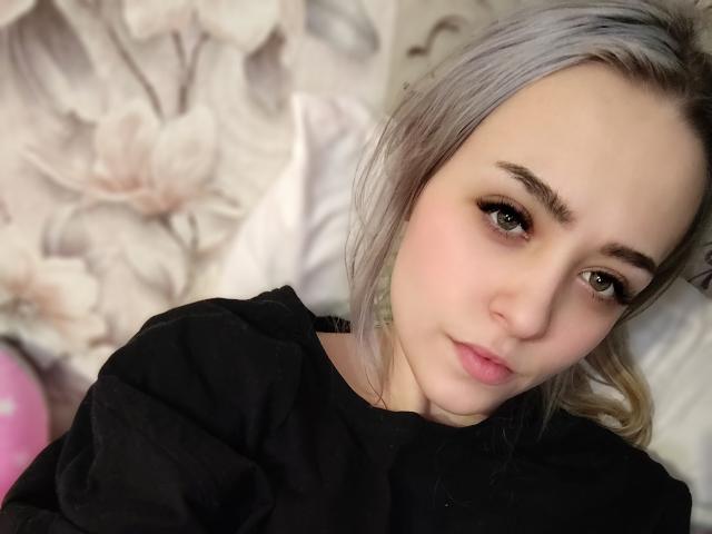 EleanorEustacia with pale skin, green eyes, and light gray hair, wearing a black shirt, lying down, looking at the camera with a subtle smile, soft pink lips.