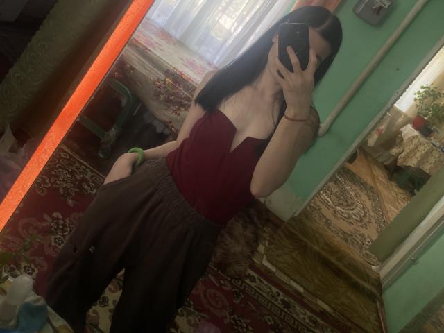 Linok with small tits, wearing a red top and brown skirt, taking a mirror selfie, her phone covering her face, in a room with colorful rugs and green walls.