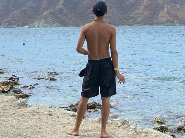 Shirtless DanteEvans with a black cap, black shorts, and a necklace stands barefoot on rocky shore, facing blue water, with mountains in the background. His ass is visible.