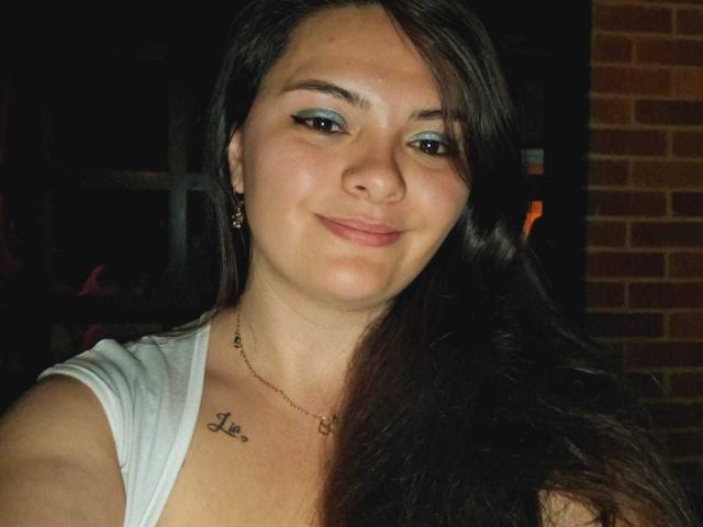 Katherin Latin tiene el pelo largo y oscuro, sombra de ojos azul claro, una nariz pequeña, un tatuaje de la palabra "Amor" en el pecho, y lleva una blusa blanca con un collar dorado..