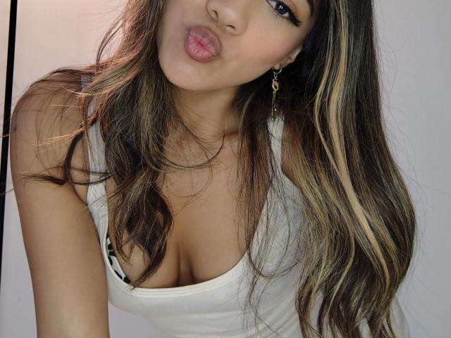 EimyRousse with puffy pink lips, long brown hair with blonde highlights, white tank top showing large tits, wearing gold earrings, looking at camera.