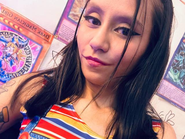 EviieGothel has brown eyes, straight black hair, pink lipstick, and a small tattoo on her right arm. She's wearing a rainbow-striped top, looking at the camera with a slight smile.