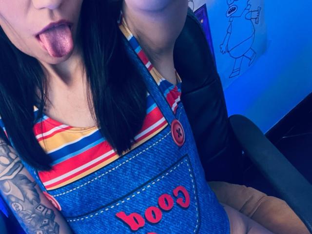 EviieGothel with black hair, tongue out, wearing striped top and denim "booo" overalls, tattoo on arm, in black chair, blue lighting. EviieGothel's tits visible, tongue pink.