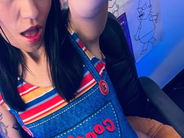 EviieGothel with black hair, red lipstick, striped shirt, denim overalls, and button, tongue out, tattoo on arm, sitting on black chair, blue light in background.