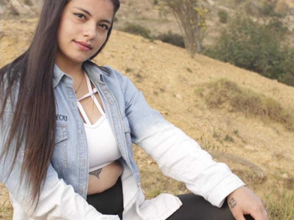 Afrodita1Hh, avec de longs cheveux noirs, portant une veste en jean bleu clair et un soutien-gorge de sport blanc, montrant un petit tatouage sous son sein droit, assise à l'extérieur..