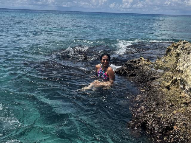 HeraDiosa with dark curly hair, wearing a colorful floral bikini, is wading in clear blue water, standing between rocky outcrops with gentle waves. She's smiling, looking happy.