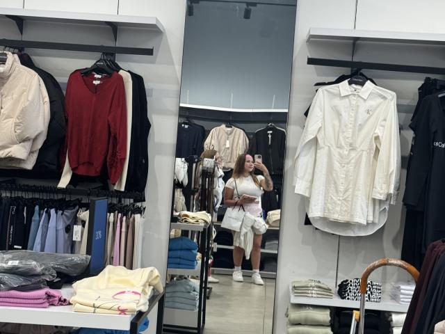 NatSorian with tits and ass visible, taking a mirror selfie in a clothing store. She's wearing a white top and pink shorts, surrounded by shirts and clothes.