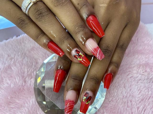 JenifferStar's hands with glossy red acrylic nails, some featuring red heart designs and white stripes, resting on a pink fuzzy surface and a clear diamond-shaped object.