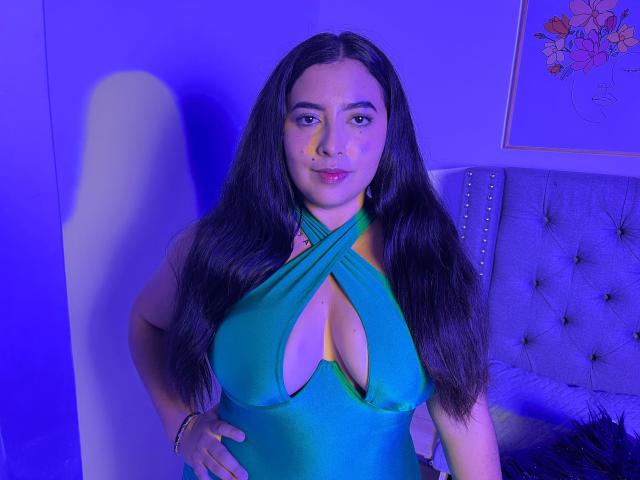 AmbarStorm con lunghi capelli castani, vestito verde a fascia, grandi seni, scollatura, in piedi, mani sui fianchi, luce blu-viola, espressione sicura, letto con testiera imbottita sullo sfondo..