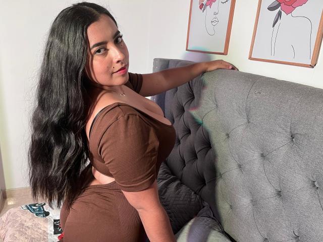 AmbarStorm with long black hair, brown off-shoulder top, and brown pants, sitting on a grey tufted sofa, looking back, showing her big ass and small tits.