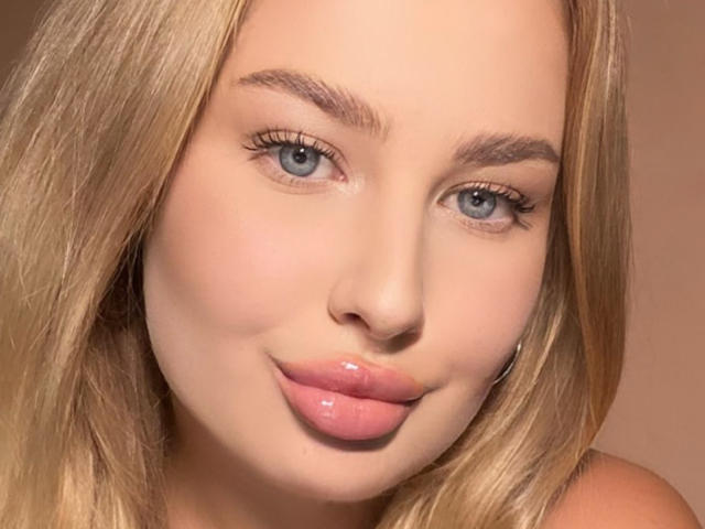 Il viso di PolinaLaur, con i capelli biondi, gli occhi azzurri e le labbra rosa lucide, è caratterizzato da un trucco leggero e un sorriso appena accennato. Le sue sopracciglia sono ben curate e la sua pelle è liscia e chiara..