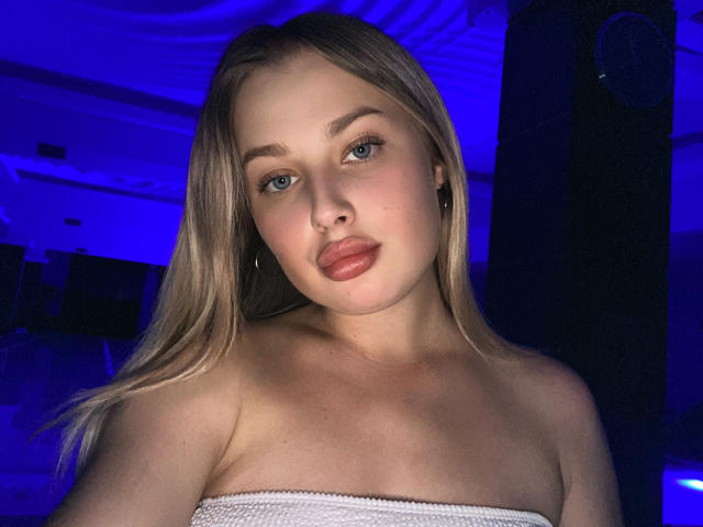 PolinaLaur, avec ses cheveux blonds, ses yeux bleus et ses lèvres pulpeuses, porte un haut blanc sans bretelles, dans une pièce éclairée en bleu. Ses épaules nues et ses petites seins sont visibles..