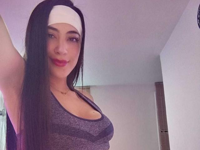 AnalisseJade has long black hair, a white headband, and a grey tank top showing her tits. She's smiling with red lipstick and a gold necklace.