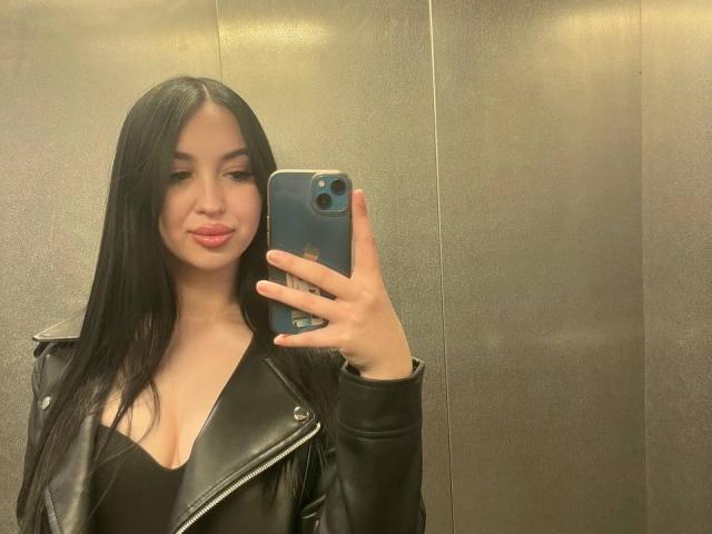 AnastaisLoveQ with long black hair, wearing a black leather jacket with a deep V-neck, holding a blue phone, taking a mirror selfie, showing her boobs.
