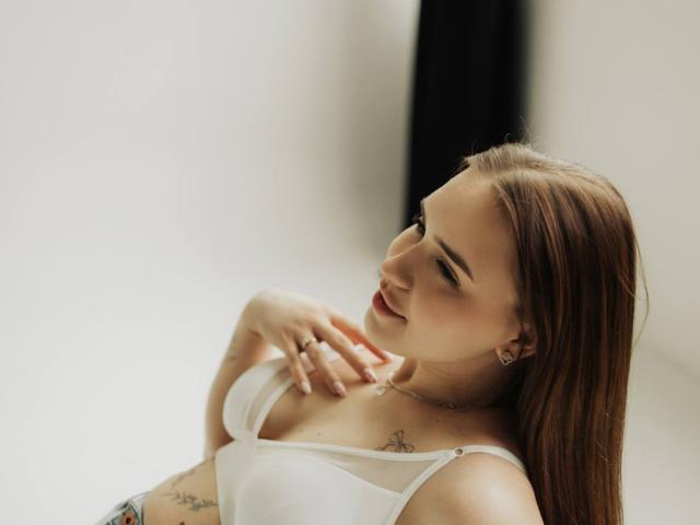 LauraHlot with light brown hair, white bra, small tattoos, smiling, hand on chest, earrings, soft light, subtle makeup, looking down, relaxed, white background, slight shadow.