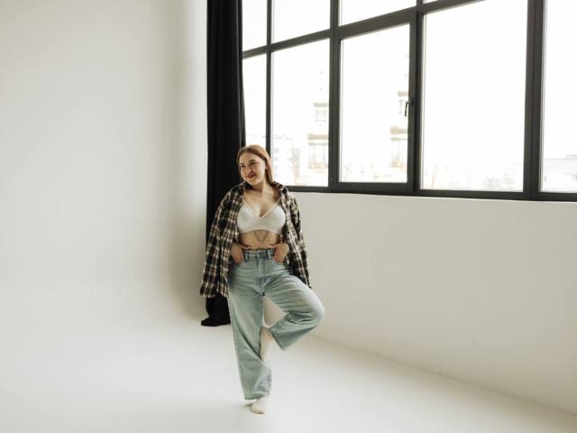 LauraHlot has small tits, wearing a white bra, blue high-waisted pants, and an open plaid shirt, standing in front of a large window, with one leg raised.