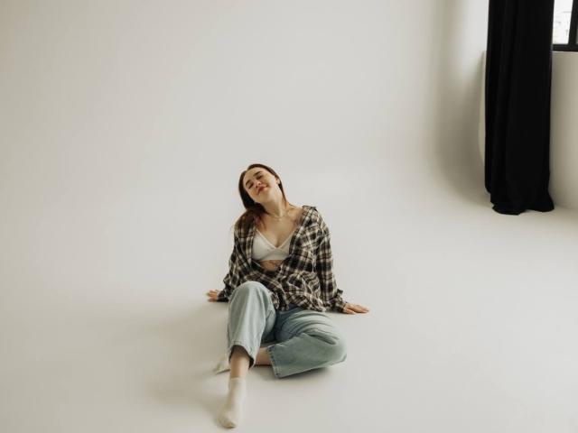 LauraHlot is sitting on the floor, wearing a plaid shirt, white bra, and blue jeans, with her head tilted back, smiling, and legs spread, showing her bare feet.