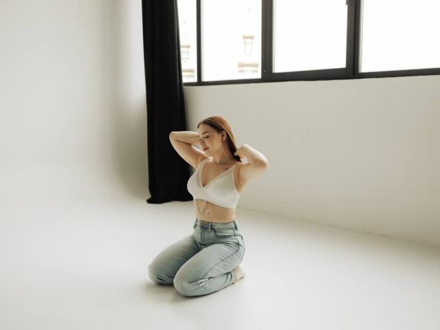 LauraHlot kneeling on white floor, arms behind head, wearing white bra and high-waisted blue jeans, showing her small tits and tattoo on right side.