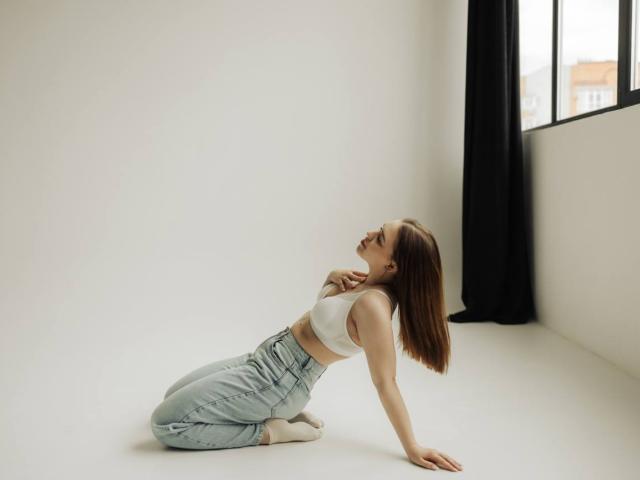 LauraHlot, kneeling, white crop top, showing small tits, light blue high-waisted jeans, brown hair, socks, arms back, head tilted up, black curtain, window, white wall, natural light.