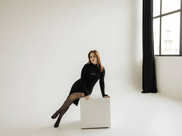 LauraHlot in a black long-sleeve dress and black tights, leaning on a white cube, with her legs crossed, wearing black heels, in a minimalist white room.