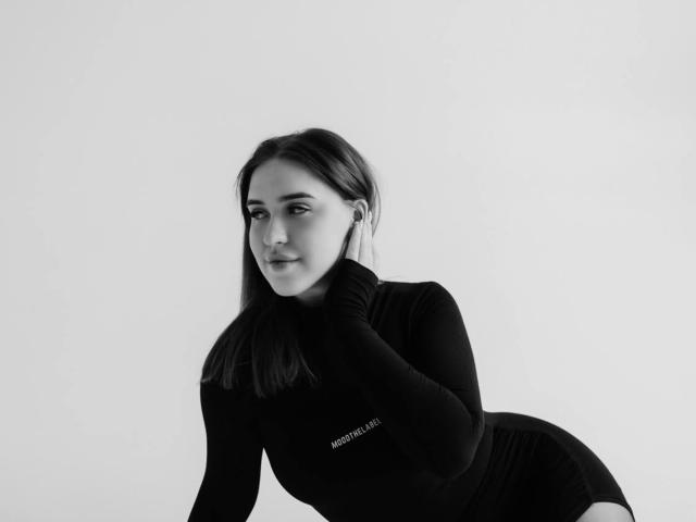 LauraHlot with straight dark hair, black turtleneck, and black shorts, leaning forward, touching her chin, showing a subtle smile, in a black-and-white photo.