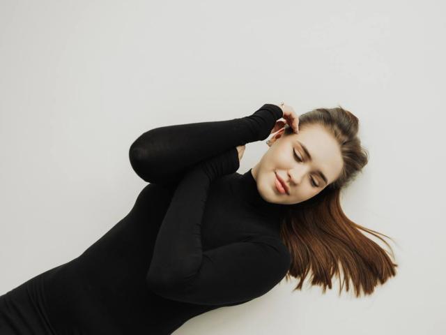 LauraHlot is lying down, wearing a black turtleneck, with her eyes closed, brown hair spread out, and one hand touching her head. She has a small smile.