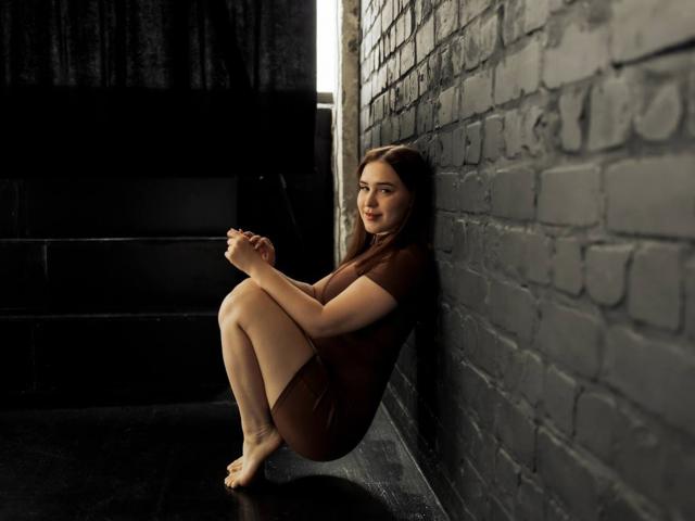 LauraHlot is sitting against a black brick wall, legs up, wearing a brown dress, hands near her face, bare feet on a black floor, looking slightly annoyed.