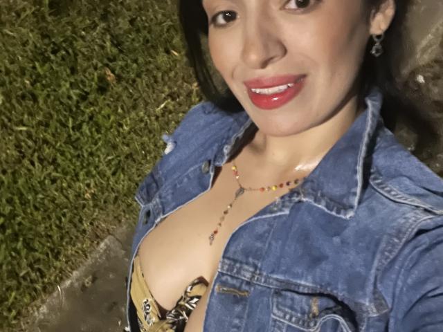 HeraDiosa with medium tits, wearing a denim jacket, red lipstick, and a beaded necklace, smiling at the camera, grass in the background.